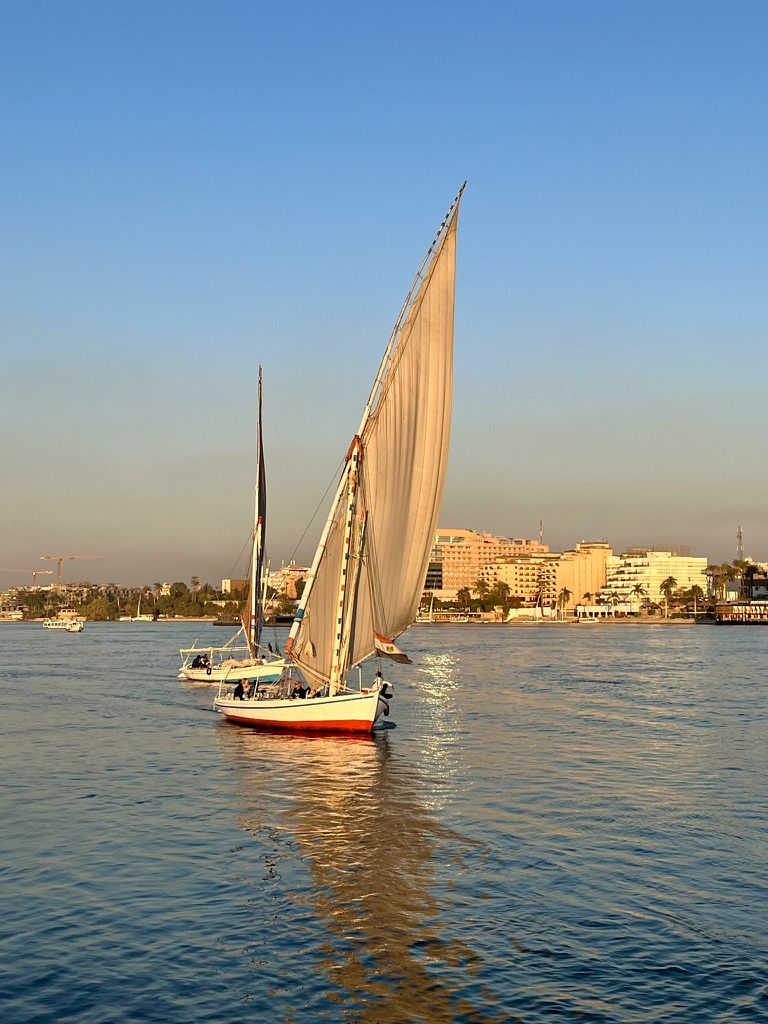 A traditional wooden felucca sailing down the Nile at sunset, silhouetted against a gradient sky of pink and orange.