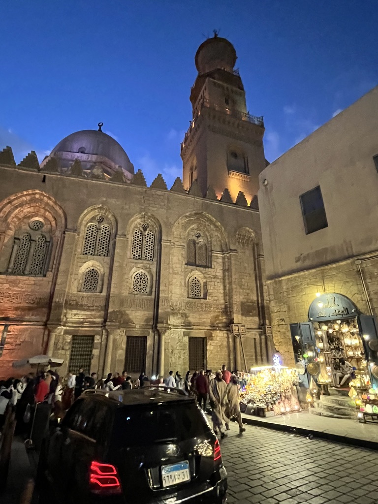 A vibrant night scene outside the Complex of Sultan al-Mansur Qalawun, where the glow of a traditional lantern shop spills onto the cobblestones of Al-Muizz Street.