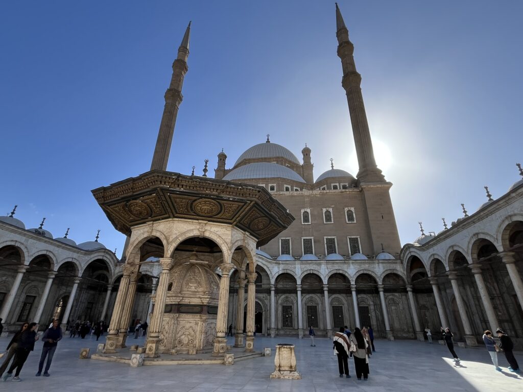The Crown of the Citadel: Sunlight breaks behind the towering minarets of the Mosque of Muhammad Ali, illuminating its grand courtyard and ornate ablution fountain.