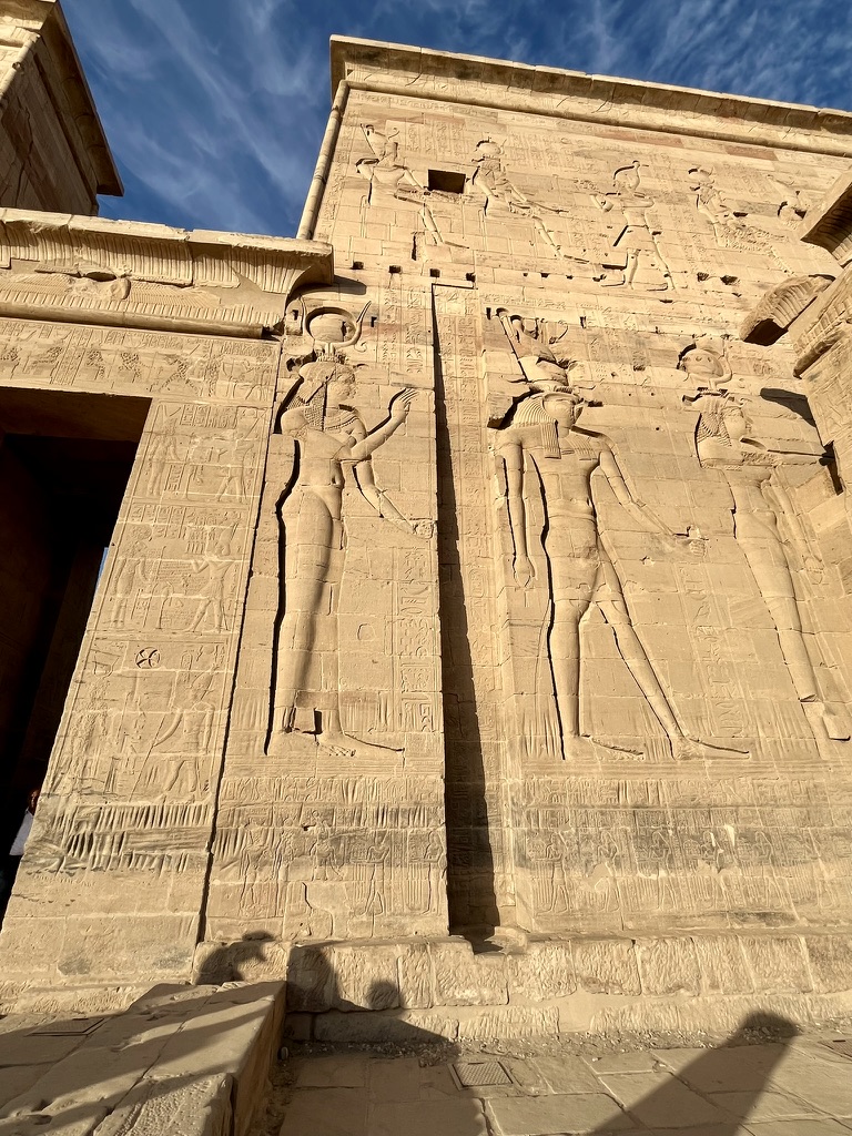 The towering First Pylon of the Temple of Isis, featuring monumental reliefs of Pharaoh Ptolemy XII Neos Dionysos smiting his enemies before the gods Isis, Horus, and Hathor.