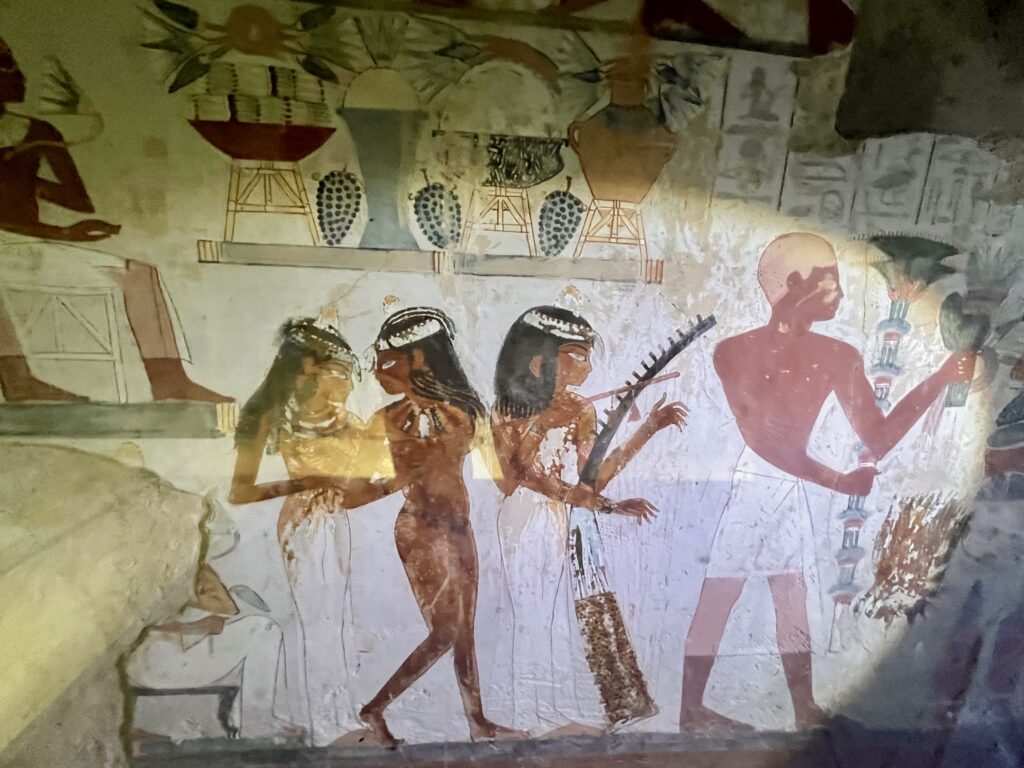 A lively banquet scene featuring three female musicians playing a harp and lute, accompanied by dancers, painted with the fluid detail typical of the 18th Dynasty.