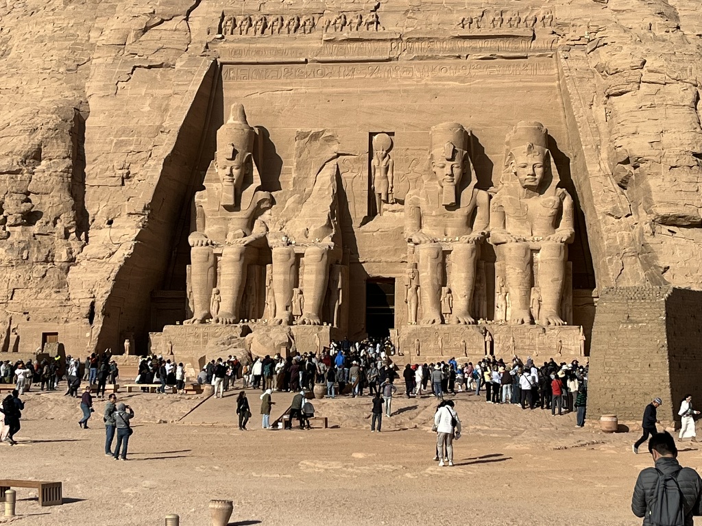 A wide view of the four colossal 20-meter-tall statues of Pharaoh Ramesses II seated on his throne, carved directly into the sandstone cliff.
