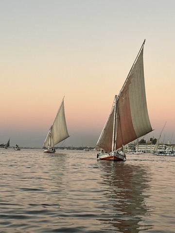 Dusk on the Nile: A wooden felucca drifting past the palm-fringed banks of Luxor as the sun sets over the Theban Hills.