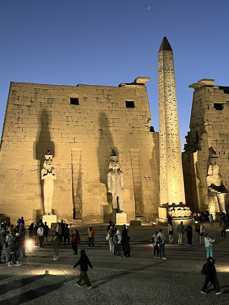 The illuminated First Pylon of Ramesses II, featuring two seated colossal statues and a single pink granite obelisk standing against the dusk sky.