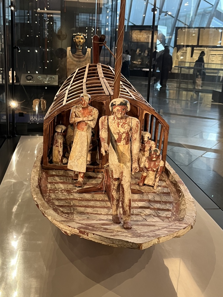 A detailed wooden model featuring a central cabin with a ribbed roof and several painted human figures, designed to accompany the deceased into the afterlife.