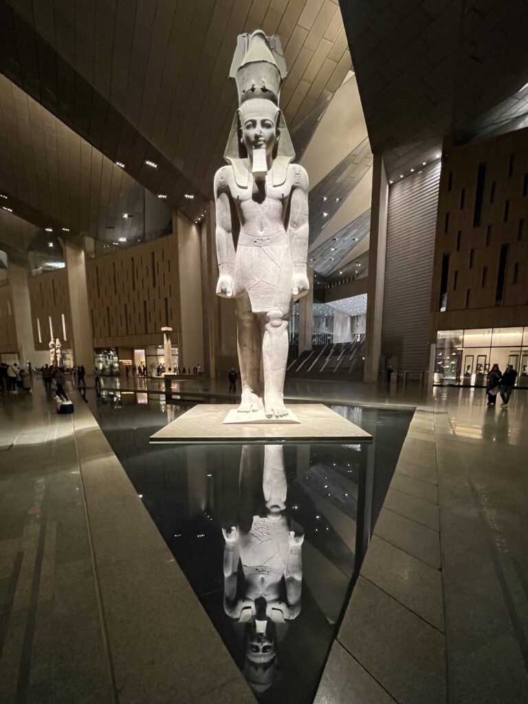 The 3,200-year-old red granite statue of Pharaoh Ramesses II, standing 11 meters tall and illuminated by the museum’s evening architectural lighting.