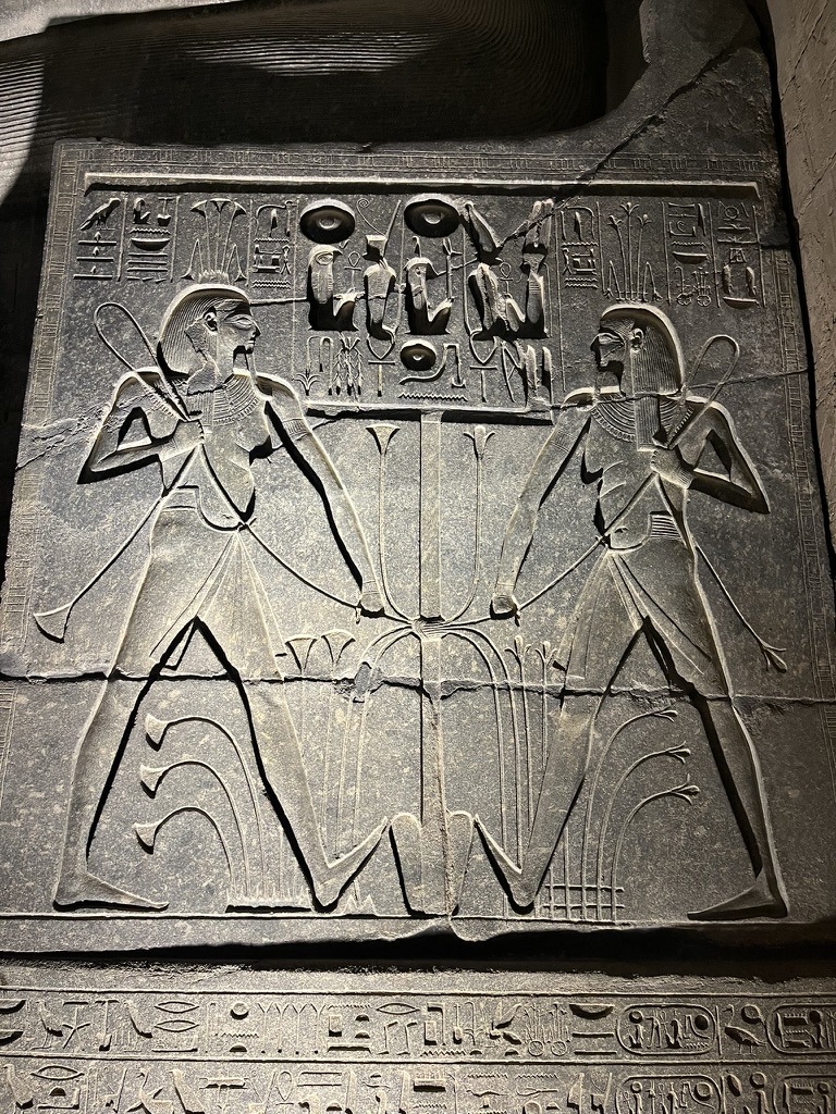 A detailed stone relief depicting two figures performing a ritual binding of the papyrus and lotus plants, symbolizing the unification of Upper and Lower Egypt.