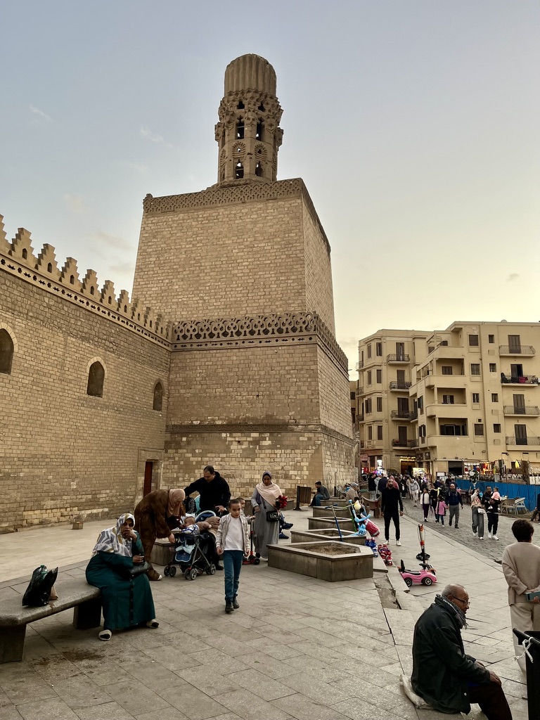 The unique flared minaret of the Al-Hakim Mosque towers over the bustling plaza near Bab al-Futuh as families gather in the evening light.