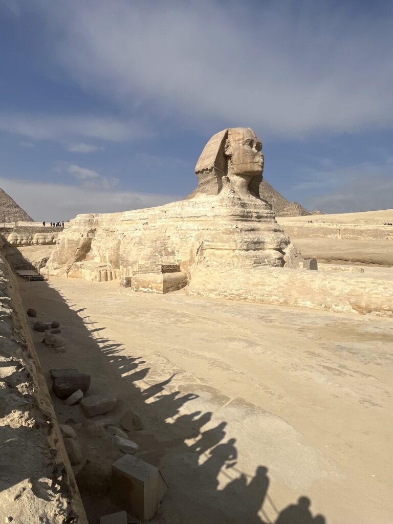The elongated shadows of visitors cast onto the limestone enclosure and the lower paws of the Great Sphinx during the noon sun.