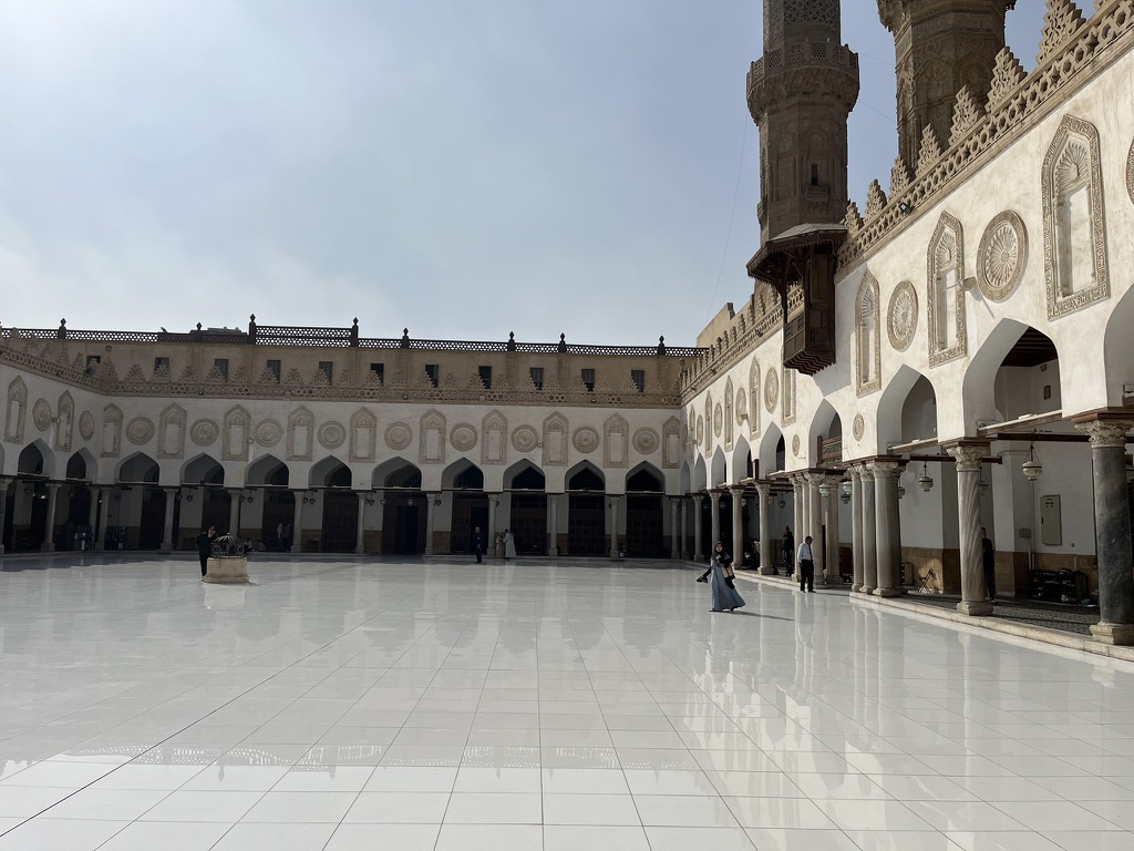 The vast, sun-drenched courtyard (Sahn) featuring a pristine white marble floor that reflects the mosque’s iconic Fatimid-era arcades and towering minarets