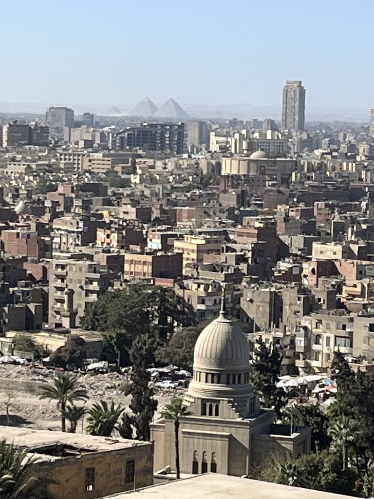 A panoramic view looking west across the Cairo skyline, where the silhouettes of the Giza Pyramids are visible on the horizon, approximately 15 kilometers away.