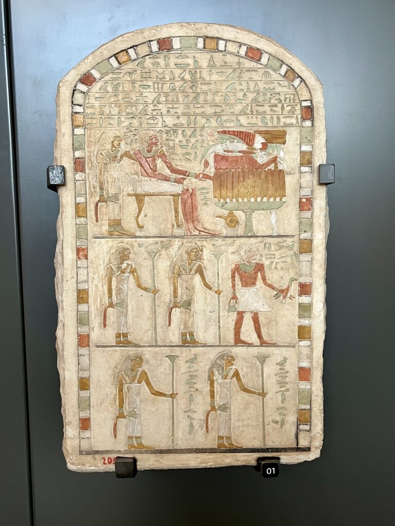 A limestone slab divided into three registers; the top shows a seated figure receiving offerings, while the lower sections depict attendants holding tall papyrus staffs.