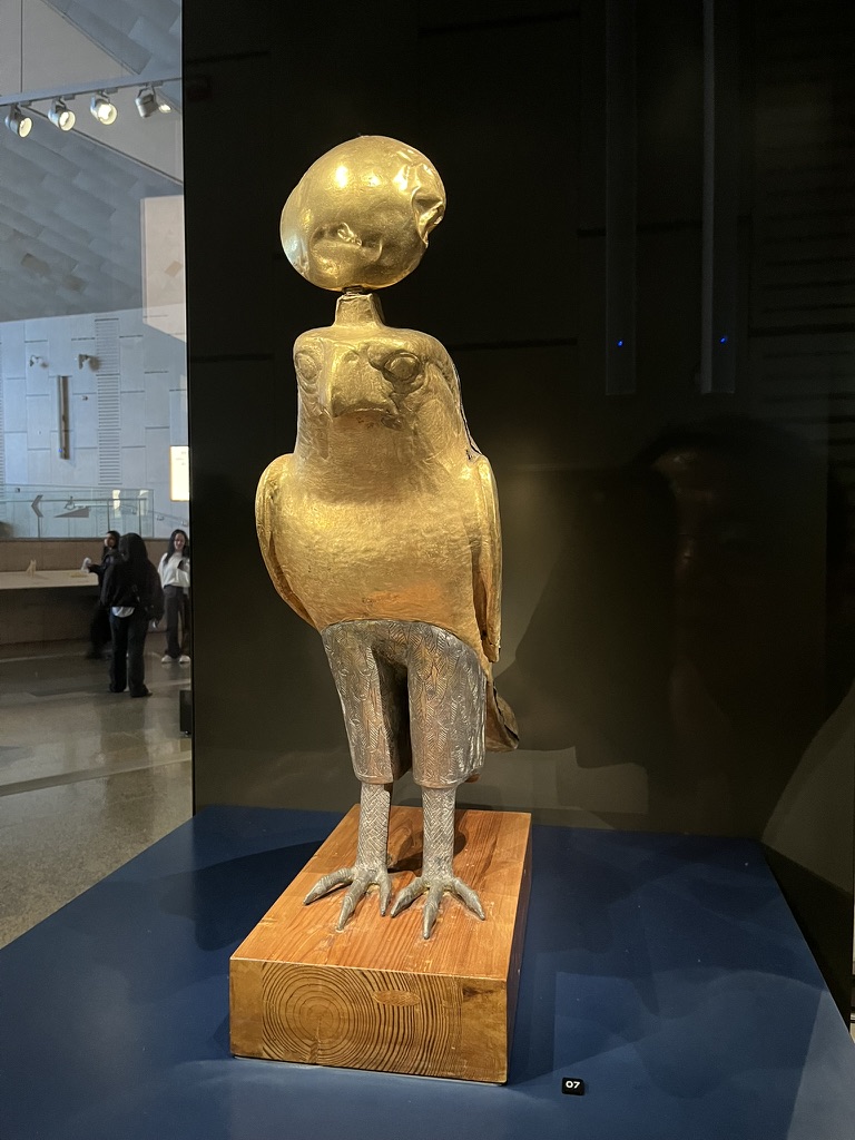 A composite metal statue of a falcon, the symbol of Horus, topped with a sun disk representing the god Ra, mounted on a modern wooden base.