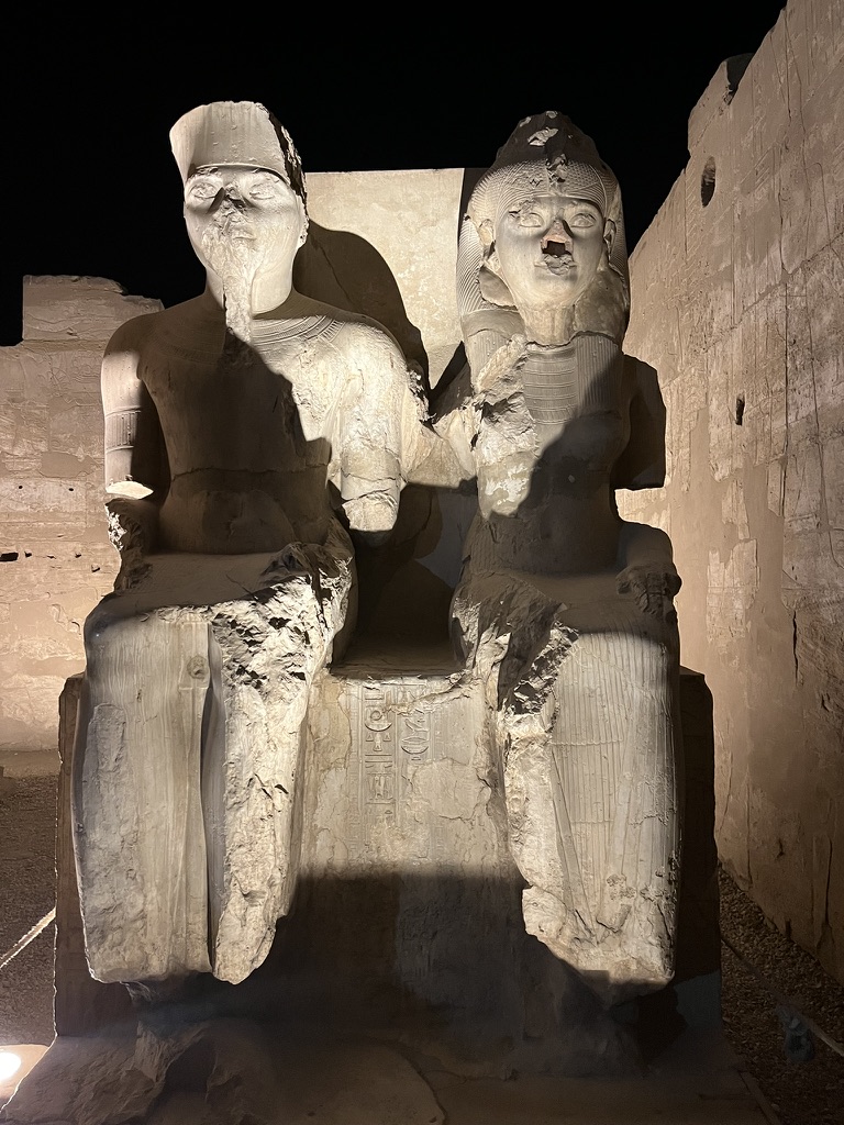A pair of weathered, seated stone statues of a royal couple, illuminated by spotlights within the temple grounds.