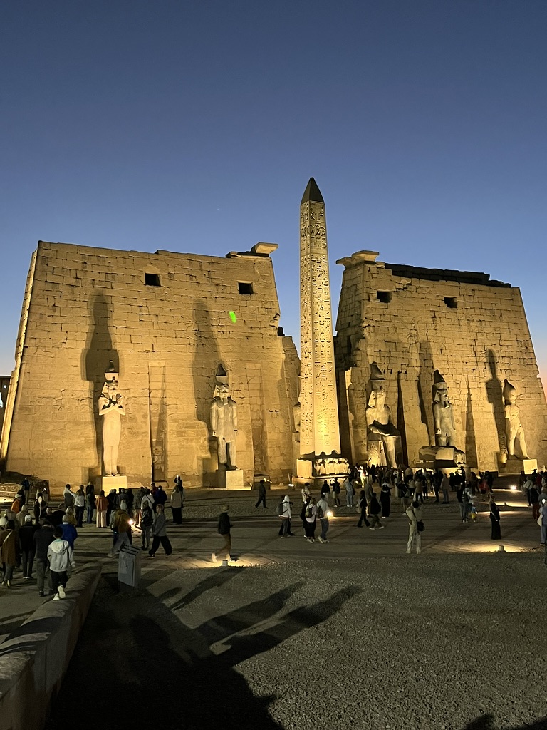 The illuminated First Pylon of Luxor Temple at dusk, featuring a single standing obelisk and colossal statues of Ramesses II.