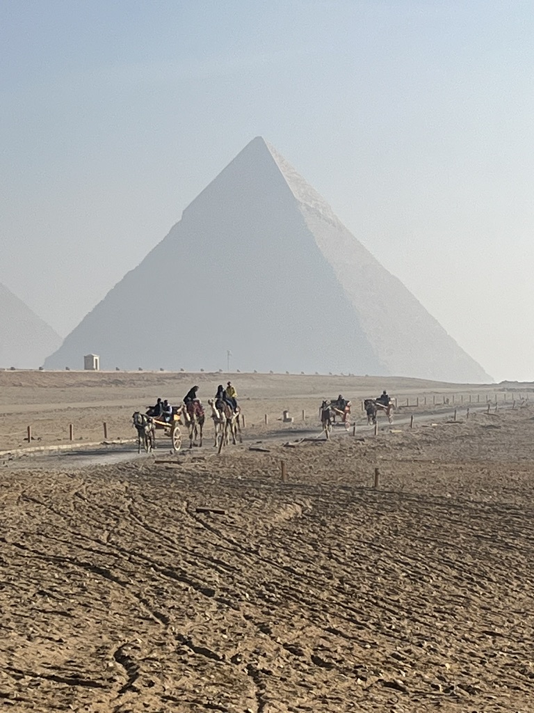 The Great Pyramid of Giza looms in the background as horse-drawn carriages and camels carry visitors across the sandy plateau.