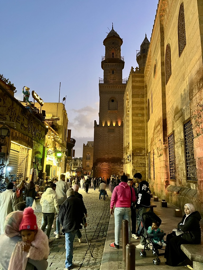 The blue hour sky settling over the massive limestone towers of Bab al-Futuh, with the warm glow of the northern end of the Khan el-Khalili market district beginning to illuminate the street below.