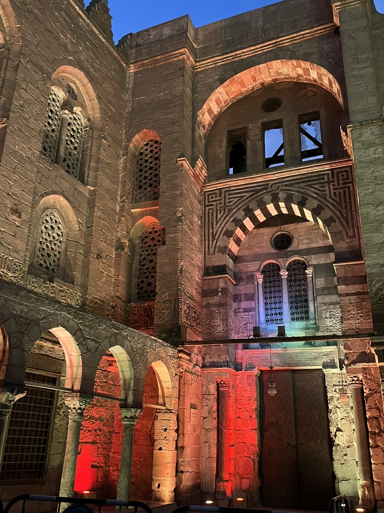 Dramatic red and blue lighting accentuates the "ablaq" (alternating stone) masonry and geometric window grilles of a historic madrasa.