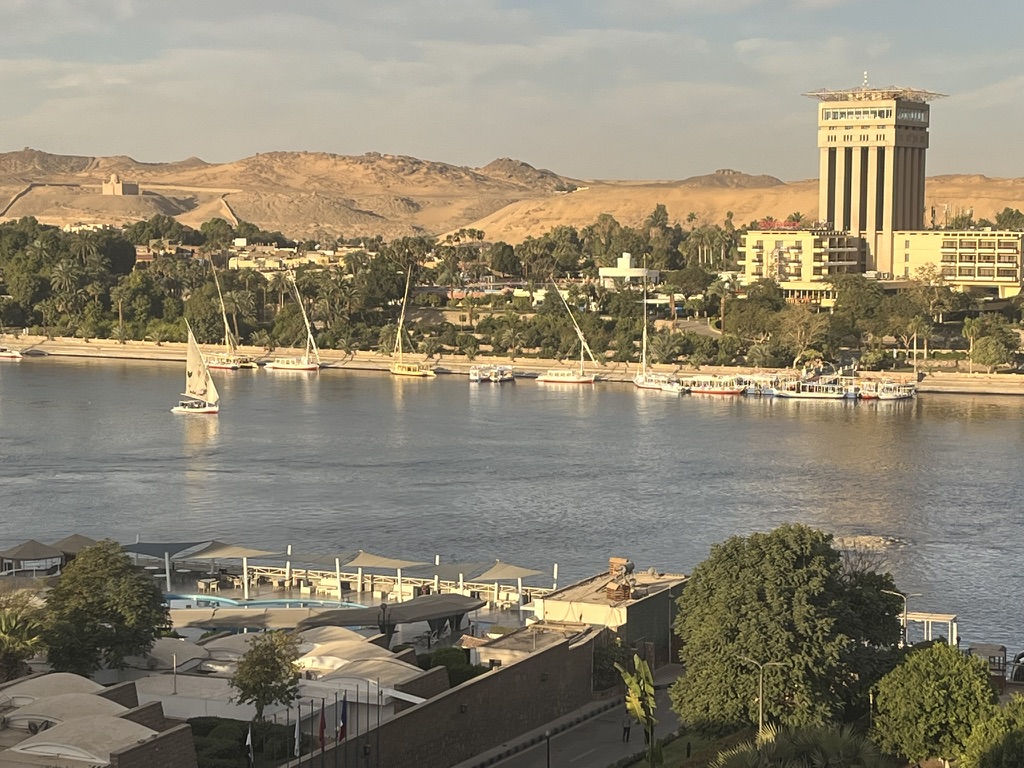 A panoramic view across the Nile looking toward the West Bank, featuring the desert hills of the Tombs of the Nobles and the sand-covered slopes of the Qubbet el-Hawa.