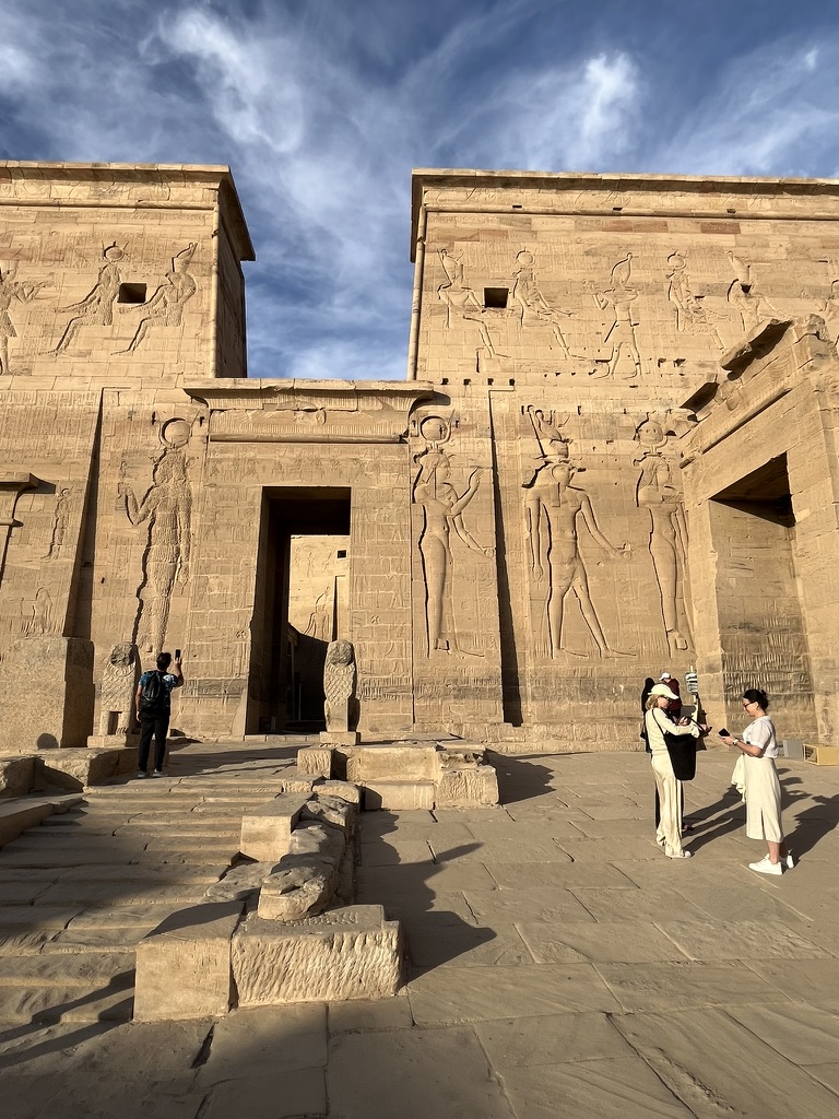 The massive First Pylon of the Temple of Isis at Philae, featuring large-scale ancient Egyptian reliefs of deities and royalty under a blue sky.